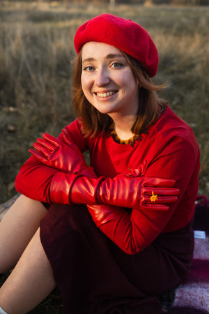 Young cute woman in red leather gloves and beret sitting on grass in autumn parkの写真素材