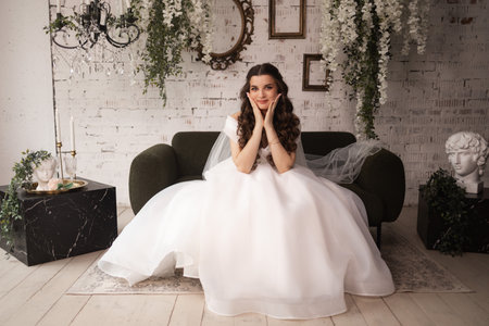 A woman in a white wedding dress sits on a couch. Concept of elegance and celebration, as the bride is dressed in her wedding gown.の写真素材