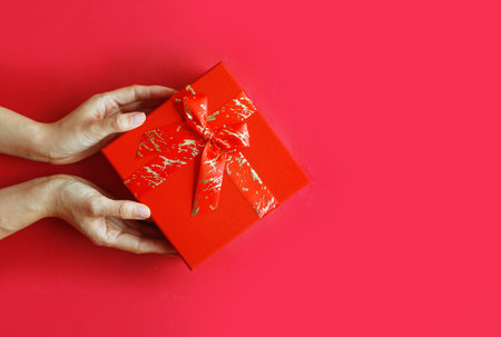 Female hands holding a red gift box on a red background. Top view. Merry Christmas and Happy Holidays greeting card, frame, banner.の写真素材