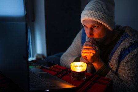 Frozen young man using laptop on sofa at home during blackout.