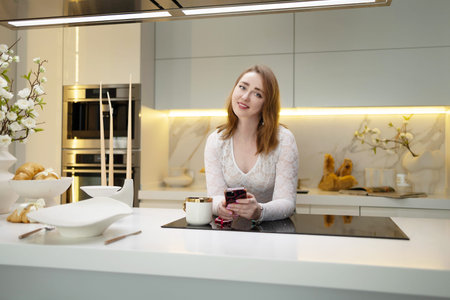 Young blonde woman enjoying a cup of coffee while using a mobile phone in a vibrant kitchen during a...