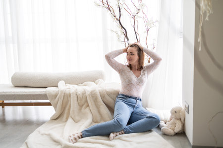 Happy woman with gentle smile and comfort at home next to big window. Young adult girl finds emotional peace in small warm gesture, relaxing in quiet moment of care and stillness.の写真素材