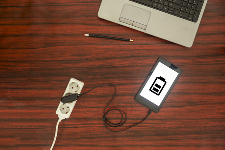 Above view of a tablet connected to electric socket on a power strip through a cable and adapter. Battery level symbol.の写真素材