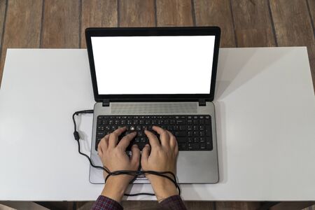 Modern slavery concept. Above view of a man hands typing on a laptop and tied with the charging cable.の写真素材