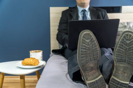 Work from home concept. Businessman dressed in a suit having a conference call on a bed with a laptop.の写真素材