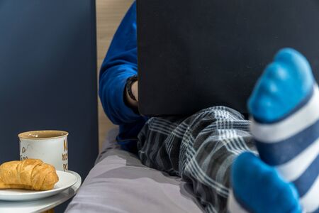 Work from home concept. Man dressed in a hoody and pijamas having a conference call on a bed with a laptop.の写真素材