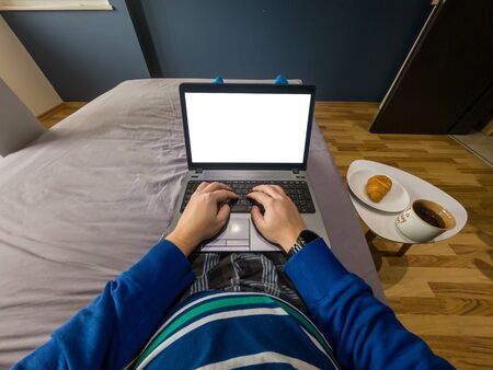Work from home concept. POV view of a man sitting on a bed with a laptop and breakfast on a small table beside the bed.の写真素材