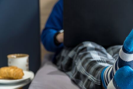 Work from home concept. Man dressed in a hoody and pijamas having a conference call on a bed with a laptop.の写真素材