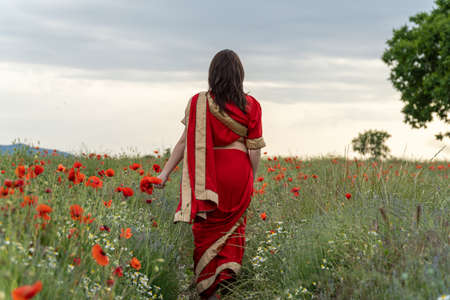 Young woman dressed in a traditional Indian costume called Sari walking in a red poppy flower fieldの写真素材