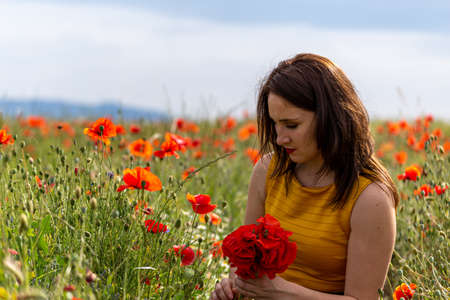 A young woman in a yellow dress in a poppy flower field with beautiful clouds in the backgroundの写真素材