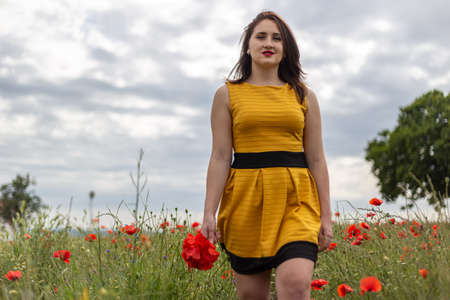 A young woman in a yellow dress in a poppy flower field with beautiful clouds in the backgroundの写真素材