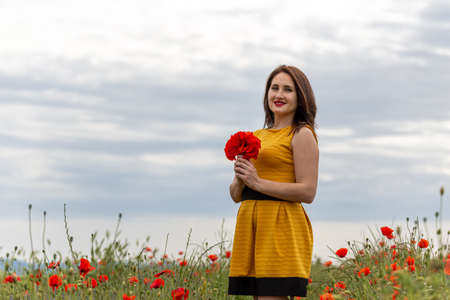 A young woman in a yellow dress in a poppy flower field with beautiful clouds in the backgroundの写真素材