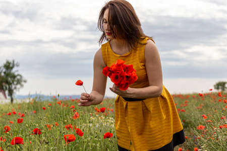 A young woman in a yellow dress in a poppy flower field with beautiful clouds in the backgroundの写真素材