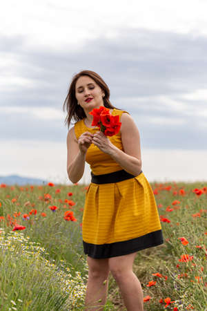 A young woman in a yellow dress in a poppy flower field with beautiful clouds in the backgroundの写真素材
