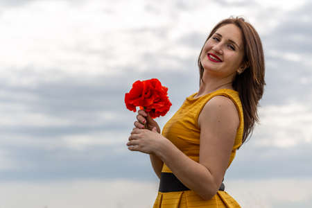 A young woman in a yellow dress in a poppy flower field with beautiful clouds in the background