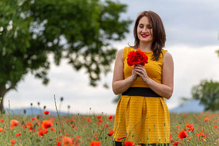 A young woman in a yellow dress in a poppy flower field with beautiful clouds in the backgroundの写真素材