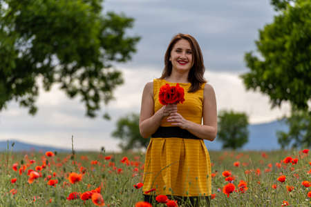 A young woman in a yellow dress in a poppy flower field with beautiful clouds in the backgroundの写真素材