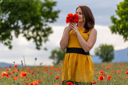 A young woman in a yellow dress in a poppy flower field with beautiful clouds in the backgroundの写真素材