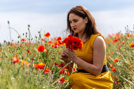 A young woman in a yellow dress in a poppy flower field with beautiful clouds in the backgroundの写真素材