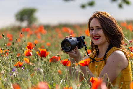 Young woman in a yellow dress holding a camera and taking pictures of a red poppy flowerの写真素材