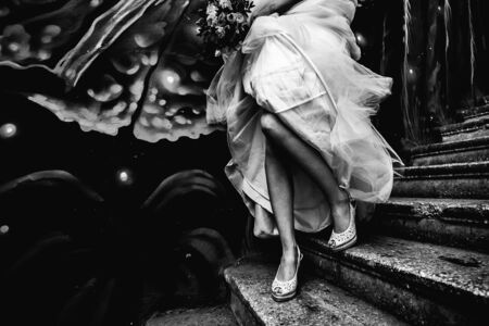 a bride in shoes runs up the stairs with a graffiti wall, holding a wedding dress in black and whiteの写真素材