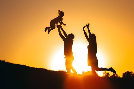 A father tosses his daughter at sunset on a hill, and next to them her mother rejoicesの写真素材