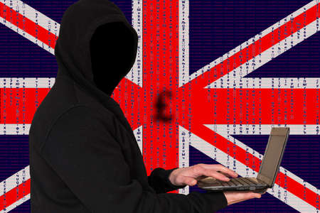 Hypothetical British hackers. An unknown person in a dark jacket with a hood holds a laptop in his...