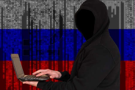 Hypothetical Russian hackers. An unknown person in a dark jacket with a hood holds a laptop in his hands against the background of the Russian flag.の写真素材