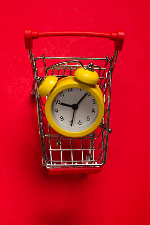 Shopping time. Yellow alarm clock in shopping basket on a red background. Copy space.の写真素材