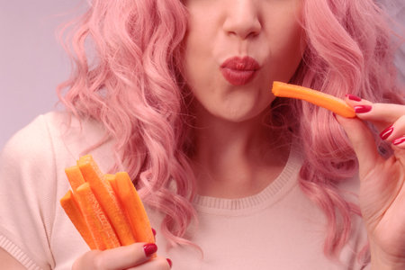 Carrots cut into strips in woman hand. Woman with pink curly hair is eating carrot close-up.の写真素材