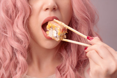Young woman with red nails and pink hair is eating sushi close up.