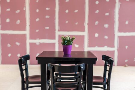 A black wooden table with a flower in a pink pot in the center and four chairs on a background of...