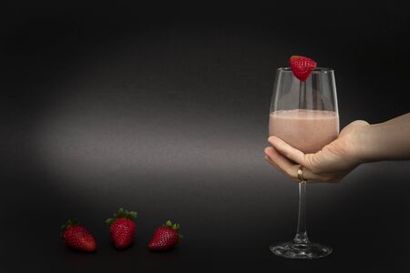 A female hand holds a vitamin cocktail in a wine glass, with one strawberry on top and three...
