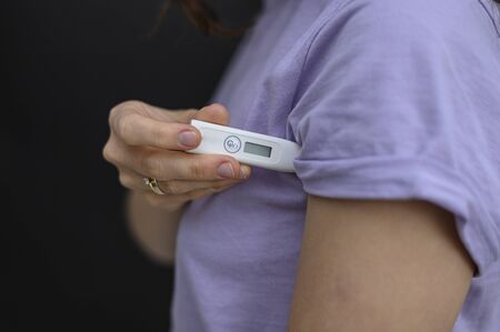 Self-monitoring and prevention during infections and pandemics. Temperature measurement is one of the important control points. The girl is going to measure the temperature, holding a thermometer under the armpit. No manicure. The thermometer is still off, it does not show anything. He is in focus. The background where the girl stands is blurred. During outbreaks of viruses, infections, flu and coronovirus, bacterial sites should be treated. Medical protective equipment is used in hospitals by doctors of various specialties, such as surgeons or nurses. Used in the professional beauty industry, cosmetology and beauty salons. Also used by sick people. If the person is a carrier of a virus or infection, he is recommended to wear a protective mask, otherwise he can infect other people. It is better for a person with a cold, cough, fever, flu to stay at home, to isolate himself or to quarantine. In an epidemic, pandemic, or environmental pollution, it is safer to carry disposable masks, gloves, and an antiseptic, that is, a disinfectant. Hand and face hygiene is important. This is a way to reduce the spread of the virus. Your safety is your health. In the midst of illness, there may be a shortage of this product, a crisis, because people buy everything in a panic. Horizontal view. Close-up. Copy space. Dark background.の写真素材