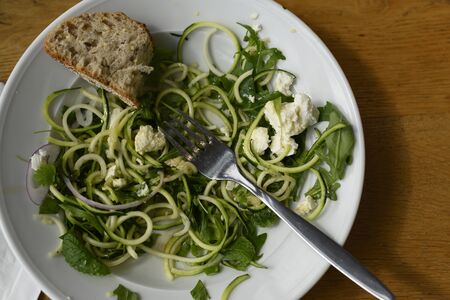 Spend free time in a cafe or restaurant, order and eat fresh, tasty and healthy food. In this photo we see a salad that can be ordered in a restaurant and it will be delicious and also very easy to make at home. The ingredients are simple, take zucchini, make pasta from it, then herbs, red onion and feta. She may be vegetarian. And it's all. We mix everything and sprinkle with fresh herbs and pour over the sauce. For example, olive oil. Bon Appetit. On this we see the half-eaten salad. Perhaps the person went to the toilet, or ate, or in a hurry. The concept of healthy and vitamin nutrition. Ideal for cafes, cafeterias and restaurants. It can serve as a motivational picture. Light composition on a wooden table. Horizontal view.の写真素材