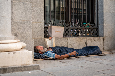 MADRID. SPAIN. 07/25/2022. A homeless man with a cigar in his hand, with a watch, lies under a blanket, reading a book on the street near the royal palace in the center of the capital.のeditorial素材
