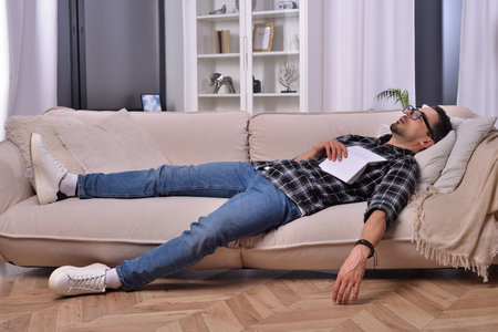 Bookworm's slumber. A man, donning stylish glasses, drifted off to sleep while indulging in a book on a comfortable couch in his inviting living room. Peaceful dreams amid the world of literature.の写真素材