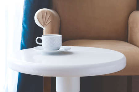 White tea cup and saucer on a round white table against a beige chairの写真素材