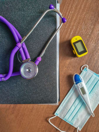 Stethoscope on a black plastic folder, thermometer, fingertip pulse oximeter and medical mask lying on a wooden table. Top view.の写真素材
