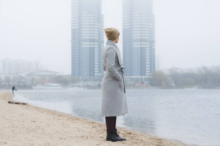 Young woman walking away alone on a river coast wearing a grey overcoatの写真素材