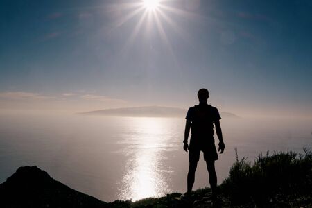 Silhouette of a happy hiker man standing on the summit. Young inspired man standing on the top of a mountain above the ocean against beautiful blue sky.の写真素材