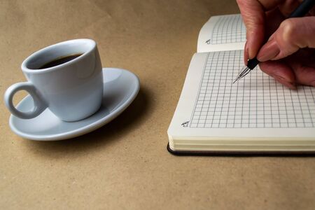 Female hand with a ballpoint pen over a notebook, next to a standing cup of coffee on a beige backgroundの写真素材