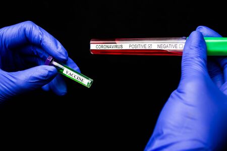 Hands in blue gloves are holding a vaccine and a test tube with blood on a coronavirus on a black backgroundの写真素材