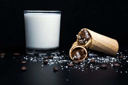 Wafer rolls with condensed milk, a glass of milk and coffee grains on a black backgroundの写真素材