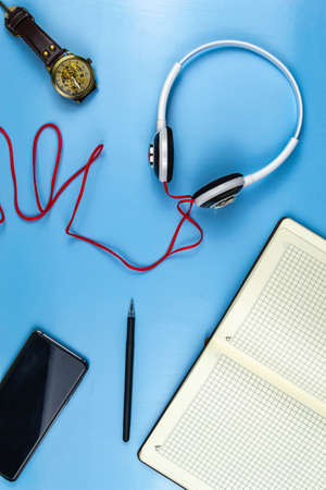 Smartphone with headphones, a cup of coffee notepad pen and glasses with a wristwatch on a blue...