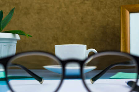 Black-rimmed glasses in the foreground. Behind glasses a cup of coffeeの写真素材