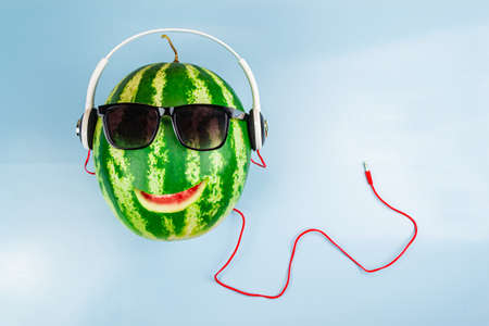 A watermelon with a cut-out smile wearing sunglasses and headphones. Blue backgroundの写真素材