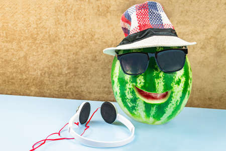 A watermelon with a cut-out smile in sunglasses and a hat, headphones next to it. Blue backgroundの写真素材