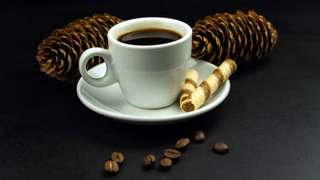 White cup of coffee, crispy striped tube, coffee grains and pine cones on black background