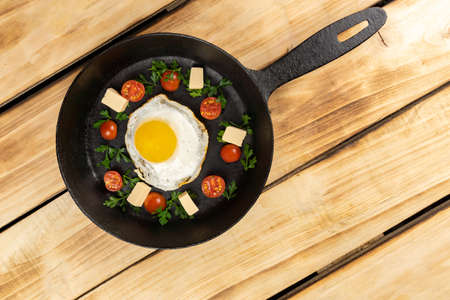 Top view of a fried egg with pieces of cheese, cherry tomatoes, herbs on a black frying pan. Wood tableの写真素材